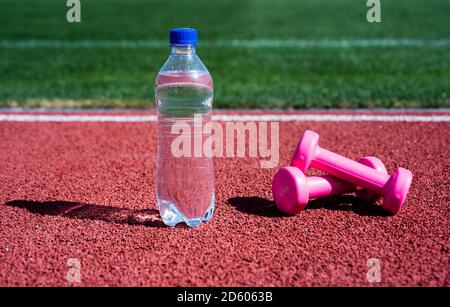 Hydratation nach Fitness. Gesunder Lebensstil. Wasserhaushalt im Körper beim Training. Langhanteln und Flasche auf Laufstrecke. Sportgerät und -Ausrüstung. Kurzhanteln auf der Rennstrecke im Stadion. Stockfoto