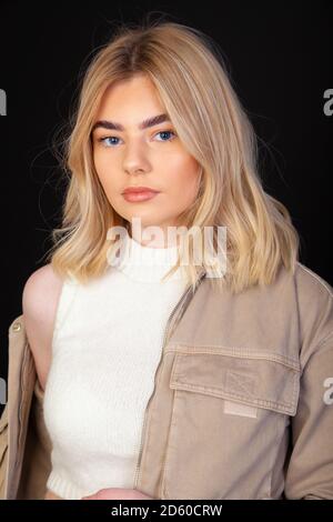 Eine schöne junge blonde Frau in ihren frühen Zwanzigern trägt eine braune Jacke und schaut direkt auf die Kamera. Stockfoto