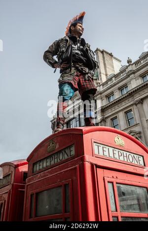 Punk-Rocker mit mohican-Haaren sitzt entspannt auf roten Telefonzellen im Zentrum von London, England, Großbritannien Stockfoto