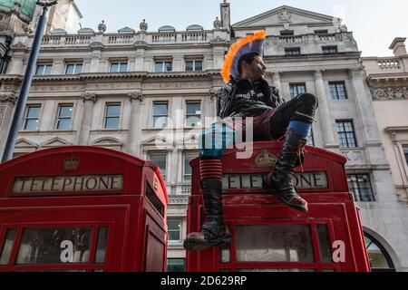 Punk-Rocker mit mohican-Haaren sitzt entspannt auf roten Telefonzellen im Zentrum von London, England, Großbritannien Stockfoto