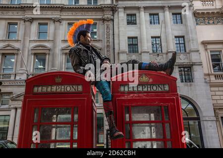 Punk-Rocker mit mohican-Haaren sitzt entspannt auf roten Telefonzellen im Zentrum von London, England, Großbritannien Stockfoto