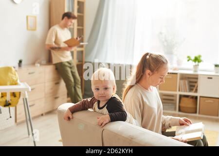Nettes Kind Blick auf Kamera, während auf dem Sofa mit sitzen Seine Mutter, die ein Buch liest und der Vater, der in arbeitet Der Hintergrund Stockfoto