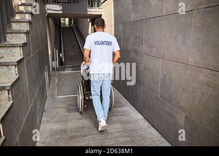 Volunteer schiebt einen Rollstuhl in einen Eingang für Behinderte Stockfoto