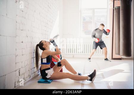 Während einer Pause im Kickboxen-Training sitzt ein junges hübsches Mädchen neben einer Wand und trinkt Wasser. Stockfoto