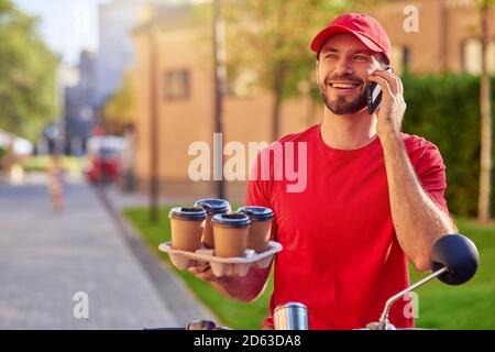 Junge lächelnde kaukasischen Kaffee Kurier am Telefon neben Roller auf der sonnigen Straße stehen. Konzept für die Lebensmittelzustellung Stockfoto