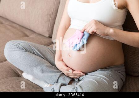 Schöne schwangere junge Frau hält ein Paar niedliche Babysocken auf ihrem Bauch und liegt auf dem Sofa zu Hause. Stockfoto