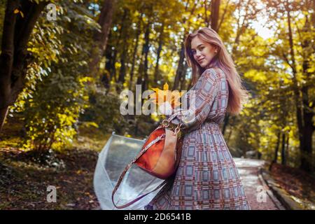 Glückliche junge Frau, die draußen mit transparentem Regenschirm nach Regen mit Blättern geht. Aktivitäten im Herbst Stockfoto