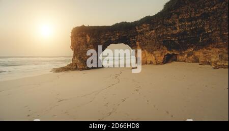 Sandstrand am Sonnenlicht mit Blick auf die Klippen. Riesiges Loch an der Felswand mit grünem Gras oben. Sumba Insel Wahrzeichen Batu Bolong, Indonesien Touristenattraktion bei niemand Naturszene Stockfoto