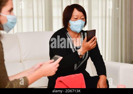 Asiatische Frau mit einer Gesichtsmaske sitzt auf einem Sofa Überprüfen Sie ihr Telefon in der Nähe einer unfokussieren Frau Stockfoto