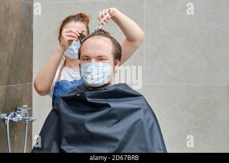 Eine Frau in medizinischer Maske schneidet einem Mann die Haare mit einer Schere, kopieren Raum Stockfoto