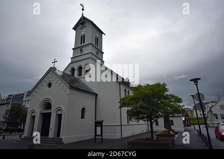 Kathedrale von Reykjavík, Dómkirkjan an einem trüben grauen Tag. Die kleine weiße Kirche hat einen Glockenturm mit einer Uhr. Bäume und Blumen säumen die Straße. Stockfoto