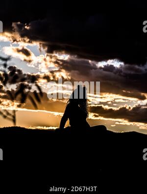 Silhouette einer Frau mit lockigen Haaren sitzt auf einem Rock mit goldener, dramatischer Wolkenlandschaft dahinter Stockfoto