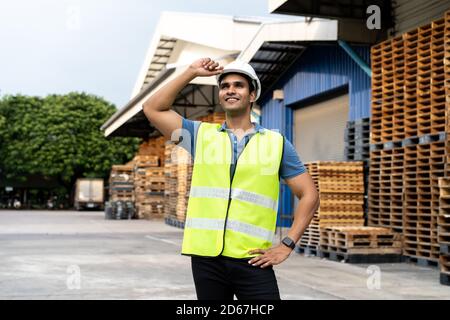 Portrait eines jungen indischen Arbeiters, der in der Logistikbranche im Freien vor dem Fabriklager arbeitet. Lächelnder, glücklicher Mann mit industriellem Hut, der weit weg die Hände auf die Hüfte im Depot schaut Stockfoto