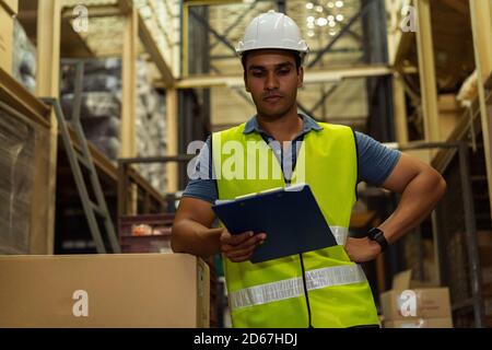 Junge indische Industriefabrik Lagerarbeiter arbeiten in der Logistikindustrie indoor. Nachdenklicher ernster Mann, der eine Zwischenablage hält und Artikel im Lager überprüft Stockfoto