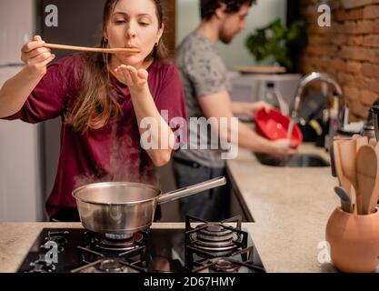 Frau, die Essen aus der Pfanne verkostet, und Mann, der Geschirr wascht Stockfoto