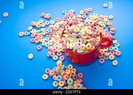 Schüssel Müsli, bunte Müsli in einer leuchtend roten Schale auf Holz, Müsli Schlaufen auf blauem Hintergrund. Schnelles Frühstück oder Snack, flat Lay. Stockfoto