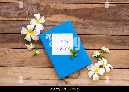 es tut mir leid, dass die Handschrift der Nachrichtenkarte auf dem blauen Tagebuch-Buch steht Mit weißen Blüten Frangipani, Jasmin Anordnung flach Lay-Stil auf Hintergrund aus Holz Stockfoto