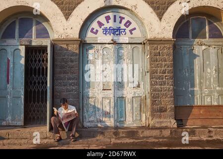 An einem frühen Morgen sitzt ein Mann vor dem (nicht mehr existierenden) Capitol Cinema, einem historischen Gebäude in Fort Area, Mumbai, Indien, in dem er eine Zeitung liest Stockfoto