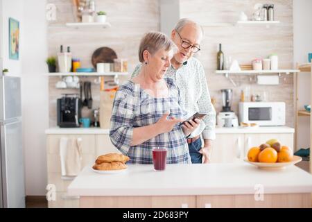 Kaukasische ältere Frau und Mann in der Küche während des Frühstücks mit Smartphone-Technologie und Internet-Anschluss. Freizeit pensionierte Frau und Mann lächeln in Domizile, entspannend. Stockfoto