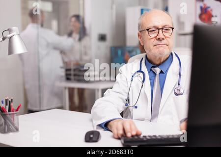 Nahaufnahme eines Arztes mit grauem Haar, das Dokument über neue Medikamente im Krankenhausschrank schreibt, und eines jungen Arztes, der mit dem Patienten im Hintergrund mit weißem Mantel spricht. Stockfoto