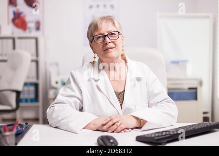 Medic mit grauen Haaren trägt Mantel in Klinik Schrank sitzen am Schreibtisch. Stockfoto