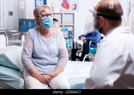 Ältere Frau diskutieren mit dem Arzt während der Konsultation im Krankenhaus Untersuchungsraum trägt Gesichtsmaske agasint covid-19. Globale Gesundheitskrise, medizinisches System während der Pandemie, kranke ältere Patienten. Stockfoto