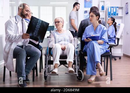 Arzt mit Stethoskop hält behinderte ältere Frau Radiographie im Rollstuhl, während mit ihr im Krankenhaus Wartebereich sprechen. Patient fragt nach seinem Termin an der Klinikrezeption. Stockfoto