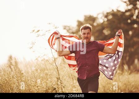 Erwachsener Mann läuft mit American Flag in den Händen draußen auf dem Feld. Fühlt Freiheit bei sonnigem Tag Stockfoto