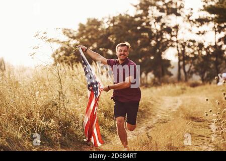 Erwachsener Mann läuft mit American Flag in den Händen draußen auf dem Feld. Fühlt Freiheit bei sonnigem Tag Stockfoto
