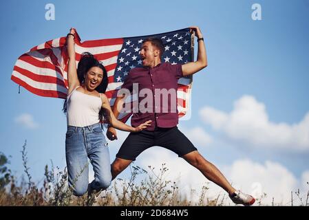 Springt hoch. Fühlt Freiheit. Schönes Paar mit amerikanischer Flagge haben eine gute Zeit im Freien auf dem Feld Stockfoto