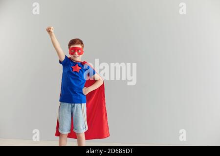 Lächelnder kleiner Junge in einem Superhelden-Kostüm, der seine Hand auf einem grauen Hintergrund hochhebt. Stockfoto