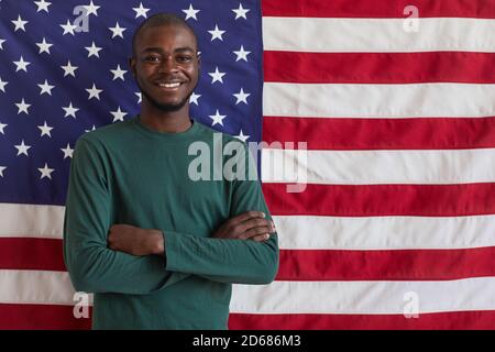 Porträt eines jungen Afro-Amerikaners, der mit gekreuzten Armen und steht Lächelnd vor der Kamera gegen die amerikanische Flagge Stockfoto
