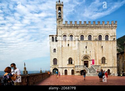 Horizontale Ansicht des Palazzo dei Consoli vor der Piazza Grande in Gubbio mit Touristen. Atemberaubender Himmel und Farben. Stadtbild. Touristen haben Masken. Stockfoto