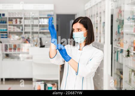 Attraktive ernsthafte Apothekerin in der Apotheke mit Maske im Gesicht Ziehen Sie blaue Handschuhe an, um zur Arbeit zu kommen Stockfoto