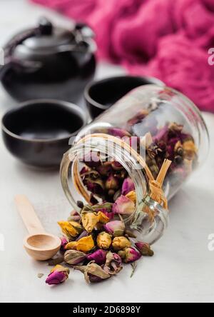 Glas Glas Rosenknospen mischen Tee mit Holzlöffel auf weißem Hintergrund mit schwarzer Teekanne und Tassen. Stockfoto