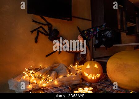 Kind Mädchen Vorbereitung Halloween in Hexenkostüm zu feiern. Große Spinne in der Hand an der Wand als Dekor. Kürbis beleuchtet und Girlande auf Tisch. Stockfoto