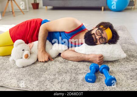 Fauler Athlet schläft friedlich auf dem Boden, umarmt einen Teddybären, mit Hanteln daneben Stockfoto