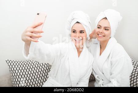 Zwei Freundinnen lachen und fotografieren am Telefon, während sie in einem Spa-Salon sitzen. Sie sind in Bademäntel gekleidet und haben Handtücher um den Kopf gewickelt Stockfoto