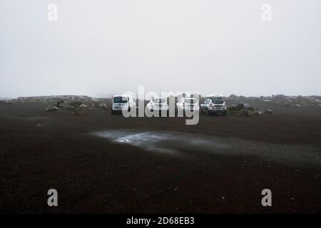 Eine Reihe von weißen touristischen Mietwagen in einem geparkt Vulkanischer schwarzer Sand Kiesparkplatz in der niedrigen Wolke Nasse Wetterlandschaft von Island Stockfoto