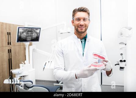 Selbstbewusster männlicher Zahnarzt steht in seiner modernen Klinik. Dentalhygieniker trägt Schutzbrille lächelnd auf die Kamera Stockfoto