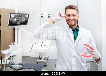 Porträt eines hübschen Zahnarztmanns, der lächelt und die Kamera anschaut und ein Zahnmodell hält. Schmerzlose Karies Behandlung in einer modernen Zahnarztpraxis Stockfoto