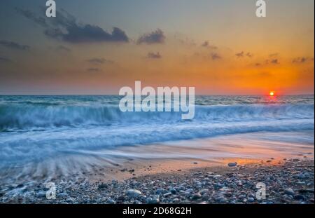 Lange Sicht auf den Sonnenuntergang und stürzende Brandung am Kieselstrand Stockfoto