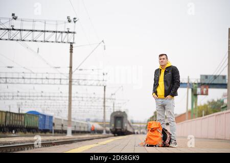 Der junge Mann steht auf dem Bahnsteig und wartet auf den Zug. Männlicher Passagier mit Rucksäcken auf Bahnsteig in Warten auf Zugfahrt. Konzept des Tourismus Stockfoto