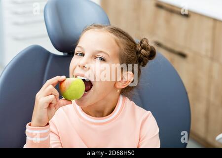 Zahnpflege für Kinder. Lächelndes Mädchen beißt einen Apfel, auf dem Zahnarztstuhl. Stockfoto