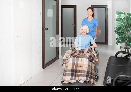 Freundliche geriatrische Krankenschwester, die ältere Frau in einem Rollstuhl im Pflegeheim-Korridor unterstützt und ältere Menschen pflegt Stockfoto