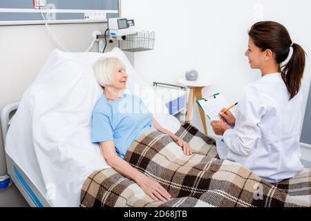 Der Arzt erklärt den Behandlungstermin, während er im Krankenhausbett eines älteren Patienten sitzt. Rehabilitation nach Herzinfarkt, Schlaganfall Stockfoto