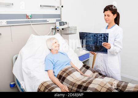 Ein erfahrener Arzt betrachtet eine Röntgenaufnahme der Wirbelsäule einer älteren Frau, während ein Patient in einem Krankenhausbett liegt. Behandlung von Erkrankungen der Wirbelsäule, Osteoch Stockfoto