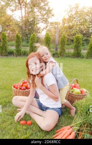 Kinder sitzen auf dem Gras und umarmen, es gibt viele reife Gemüse in der Nähe. Tomaten, Karotten, Paprika. Stockfoto