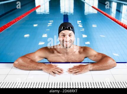 Positive Schwimmer, trägt Schwimmbrille und Badekappe, entspannen im Pool nach dem Training. Mann, der am Pool posiert Stockfoto
