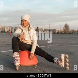 Ein Mädchen in Rollschuhe sitzt auf einer Parkplatz Hemisphäre. Eine junge Eiskunstläuferin ruht. Herbstabend. Das Mädchen trägt einen weißen Hut, Pullover, Jacke und schwarze Leggings. Speicherplatz kopieren. Stockfoto